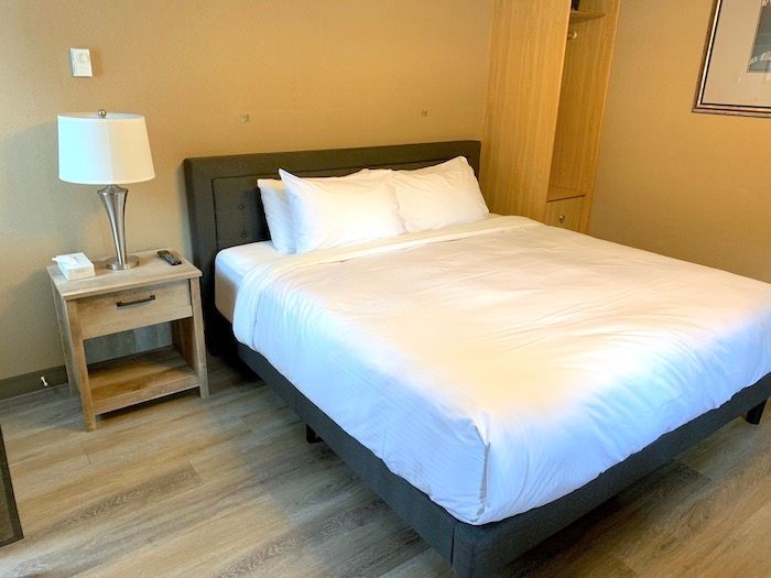 Hotel room with a queen-sized bed, nightstand with lamp, and wardrobe. Neutral colors, wooden floor.