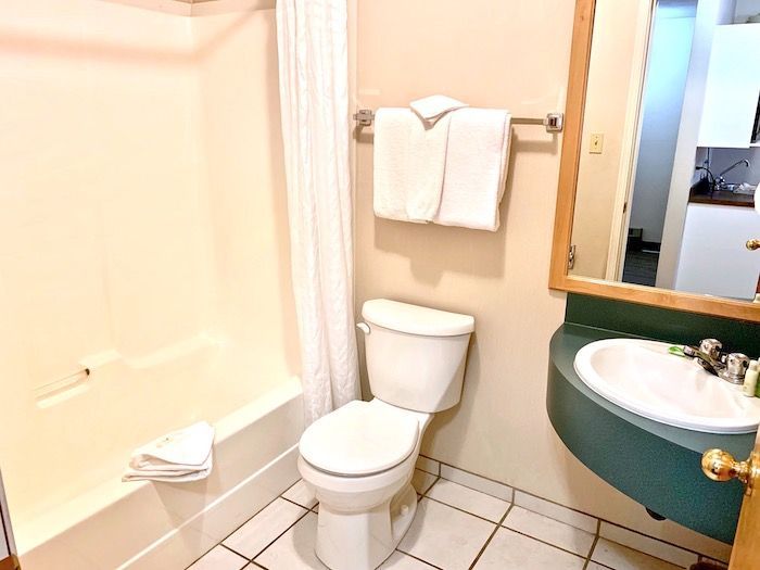 Bathroom with a toilet, sink, tub, and white towels. Beige walls, green sink surround, and a mirror.