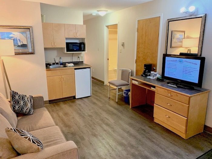 Small hotel room with kitchenette, sofa, desk, and TV; light wood cabinets and flooring.