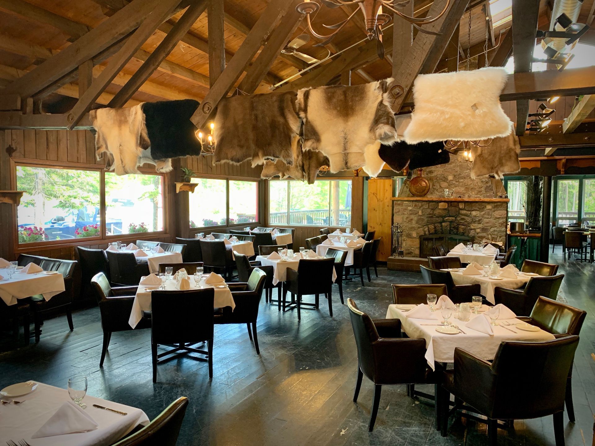 Restaurant interior: tables set with white tablecloths, dark chairs, rustic wooden beams, and animal hides hanging.