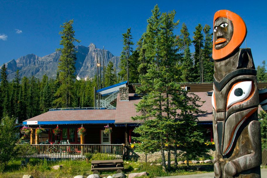 A totem pole stands in front of a lodge, with mountains in the background under a blue sky.