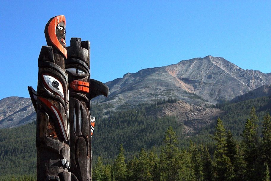 Two totem poles with mountain backdrop and blue sky.