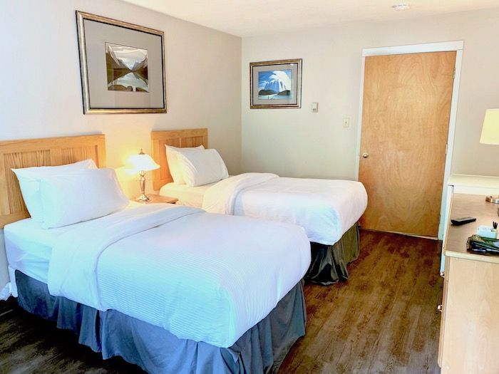 Two twin beds in a hotel room with art on the walls, a wooden door, and a nightstand.