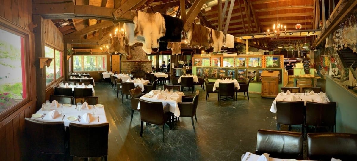 Interior of a rustic restaurant with wooden beams, tables with white tablecloths, and animal hides hanging from the ceiling.