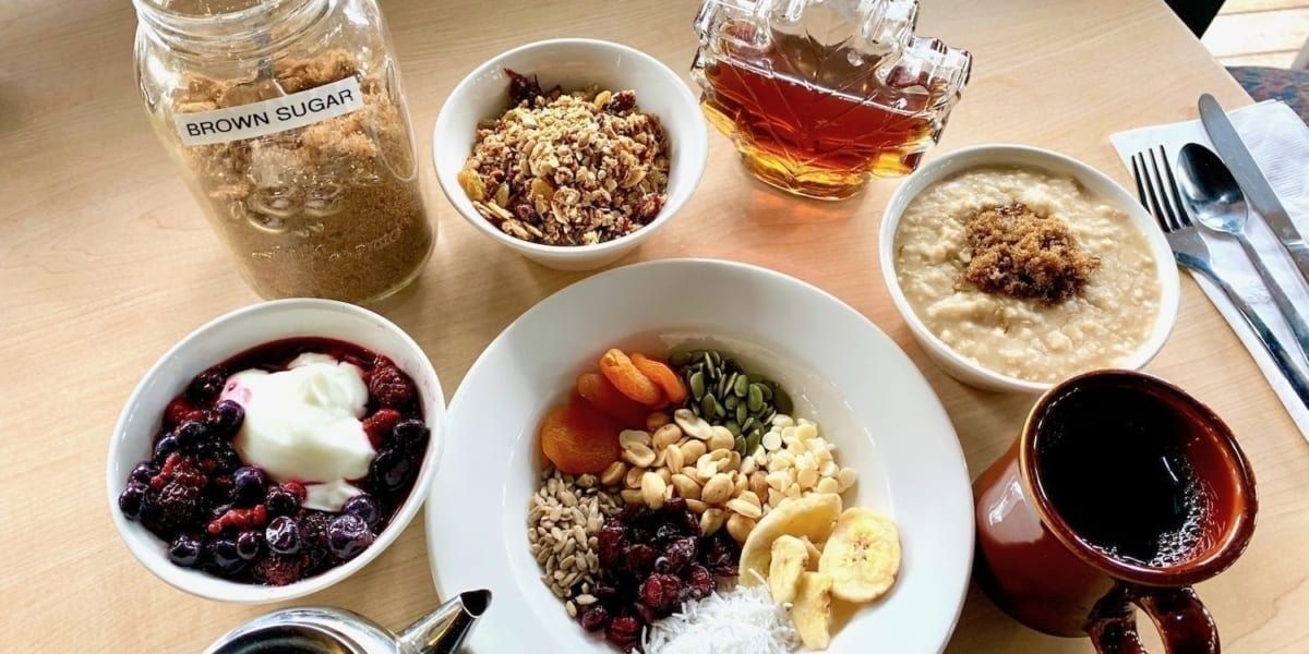 Breakfast spread with oatmeal, berries, fruit, granola, syrup, coffee, and a jar of sugar.