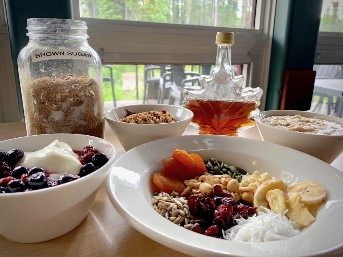 Bowls of oatmeal and toppings, maple syrup, granola jar, and a window.