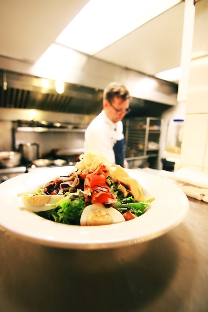 Salad on a plate in focus, chef in blurred background of a commercial kitchen.