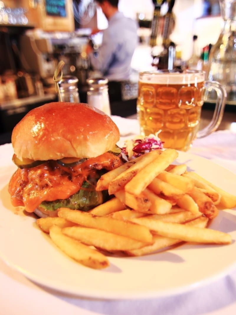 Burger, fries, and beer on a white plate in a restaurant. A person is in the blurred background.