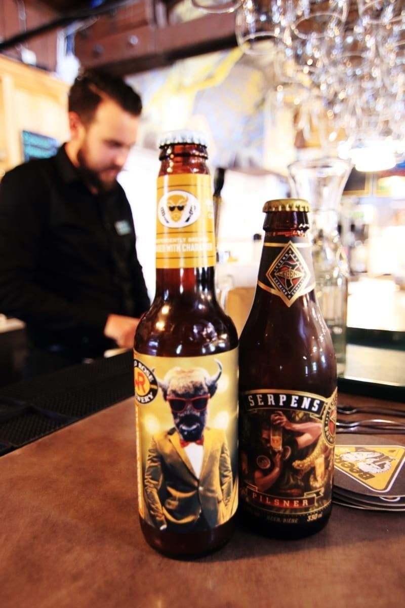 Two beer bottles on a bar, a bartender in background. One bottle has a person wearing a suit on the label.
