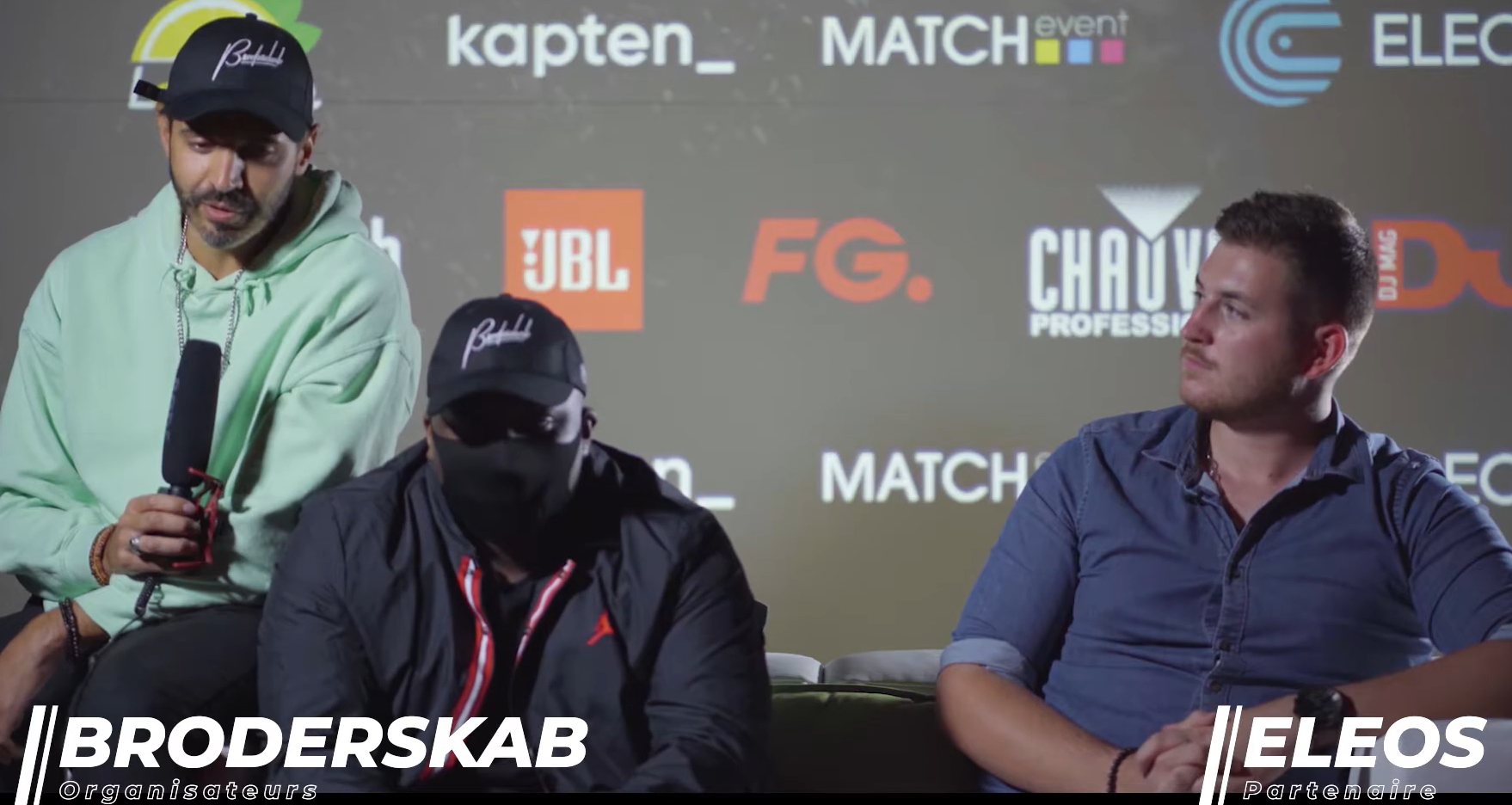 Trois personnes sont assises devant un fond portant une marque ; la personne au centre porte un masque noir, tandis que celle de droite regarde.