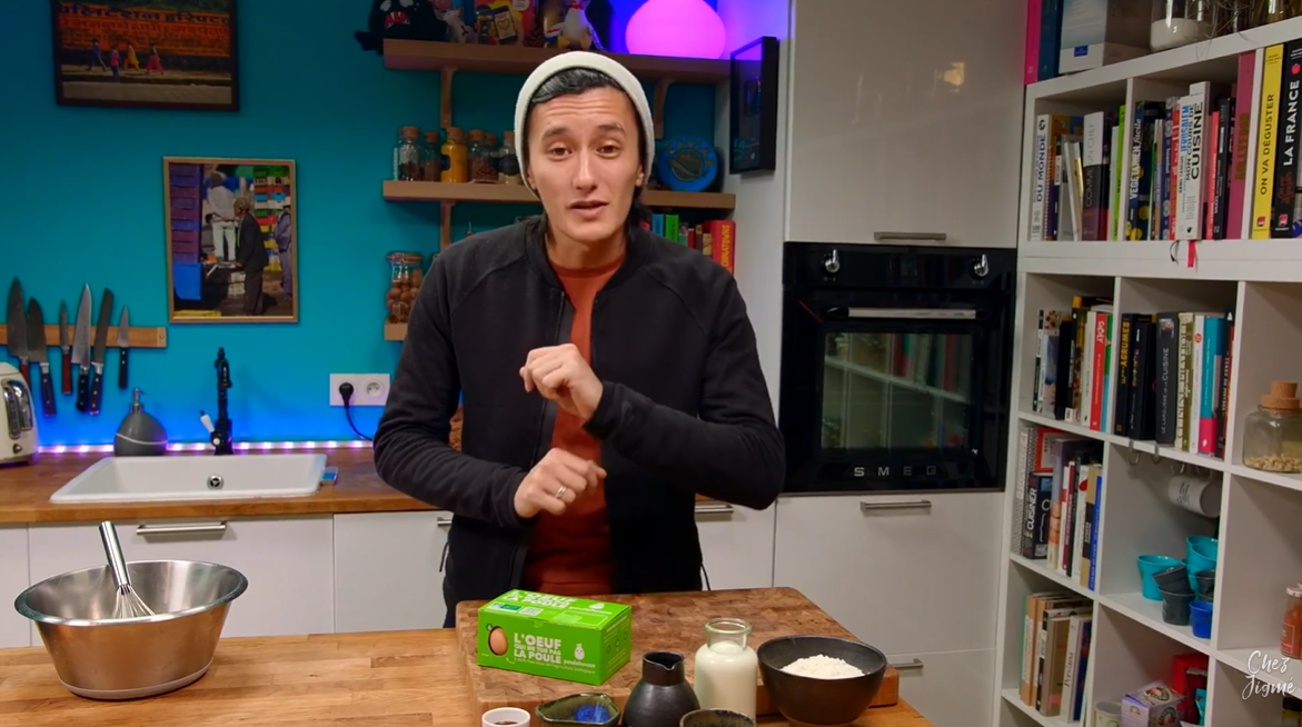 Une personne portant un bonnet et une veste présente un paquet vert sur un comptoir de cuisine en bois, avec des étagères et un four derrière.