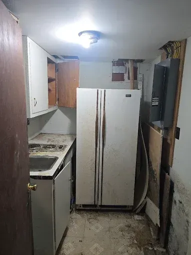 Dirty, small kitchen with a refrigerator, cabinets, and a sink. Debris covers the floor and exposed walls.