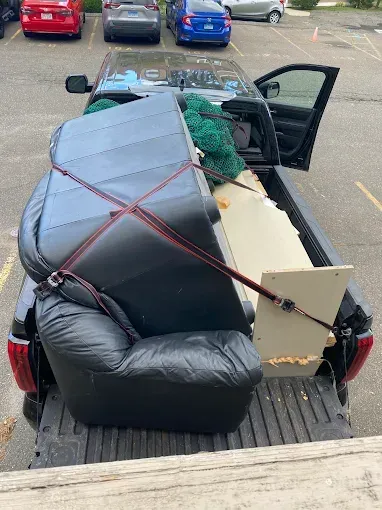 A black pickup truck bed is packed with furniture, including a large black recliner, secured with straps.