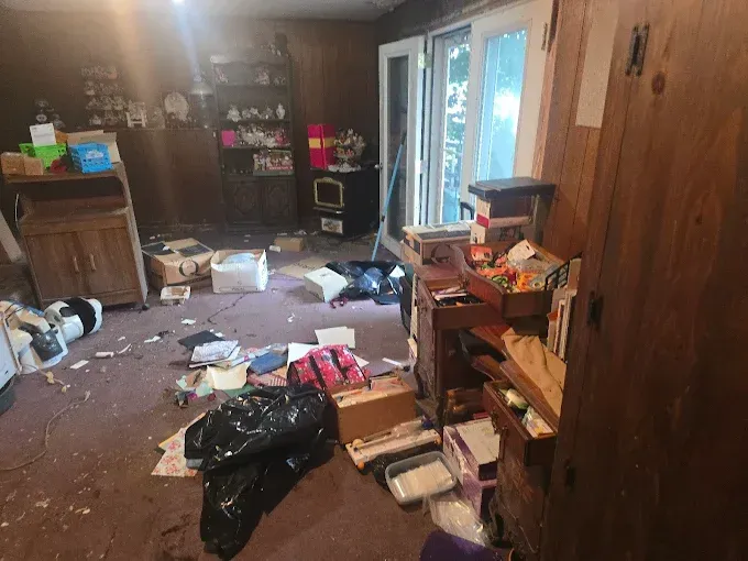 Cluttered room with debris on the floor, boxes, and furniture; door open to outdoors.