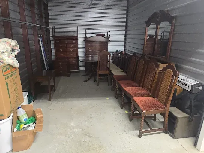 A storage unit filled with antique furniture: chairs, dresser, mirror, and table.