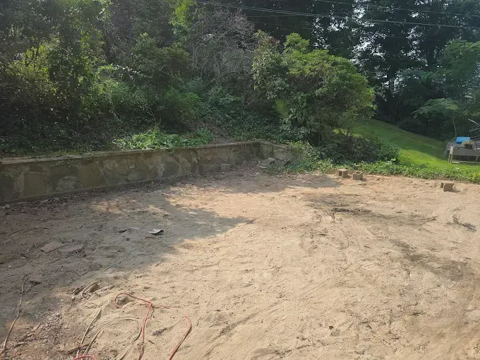 Dirt lot with concrete wall and overgrown hillside. A car is visible on the right.