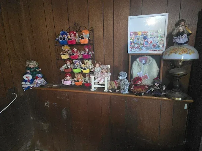 Shelf with doll collection, framed art, and a lamp against wood paneling.