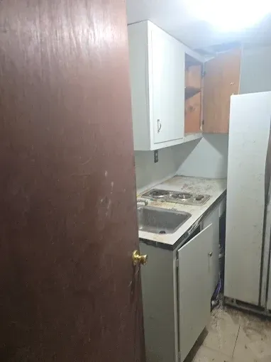 Small, outdated kitchen with white cabinets, stove, sink, and a fridge; door is partially visible on the left.