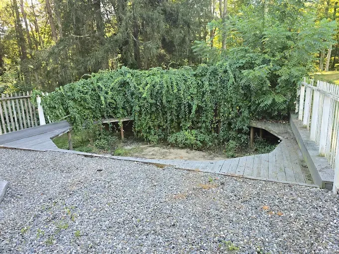 Gravel path leads to a wooden deck with overgrown green bushes and small benches, bordered by a white picket fence.