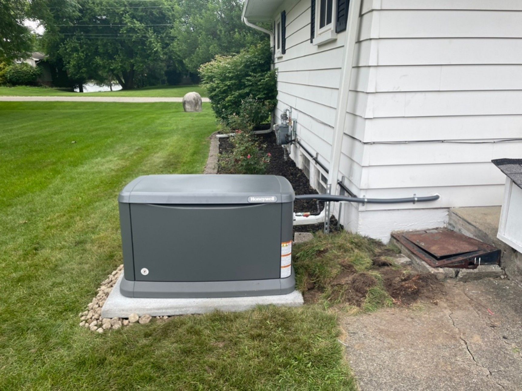 A grey standby generator sits on a concrete pad next to the white siding of a house, connected by conduit.