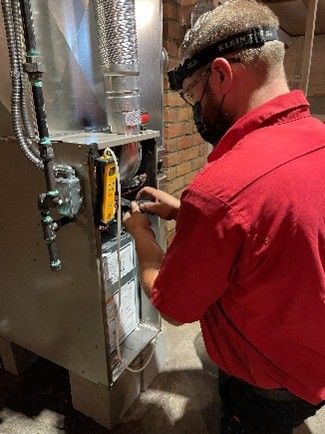 HVAC technician working on a furnace in a brick-walled room. He wears glasses, a mask, and a headlamp.