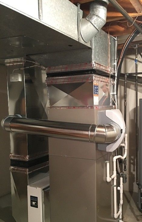 Metal HVAC ductwork with rectangular and cylindrical components against a light-colored wall.