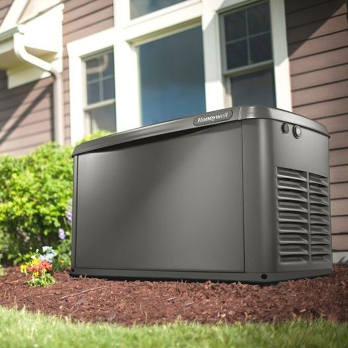 A dark grey Honeywell standby home generator sitting on a mulch bed next to the side of a house.
