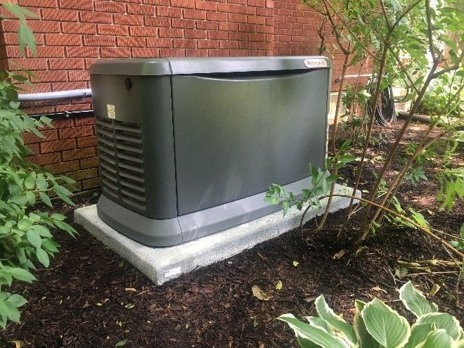 A dark-colored standby generator on a concrete pad near a brick wall and foliage.