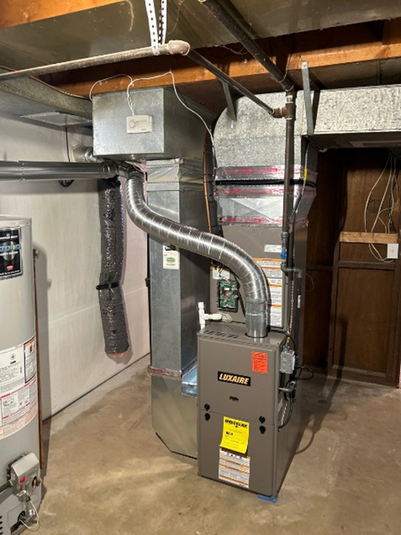 A gray Comfortmaker furnace sits in a basement with silver metal ductwork and a nearby water heater.