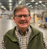 A man wearing glasses and a green vest is smiling in a warehouse.