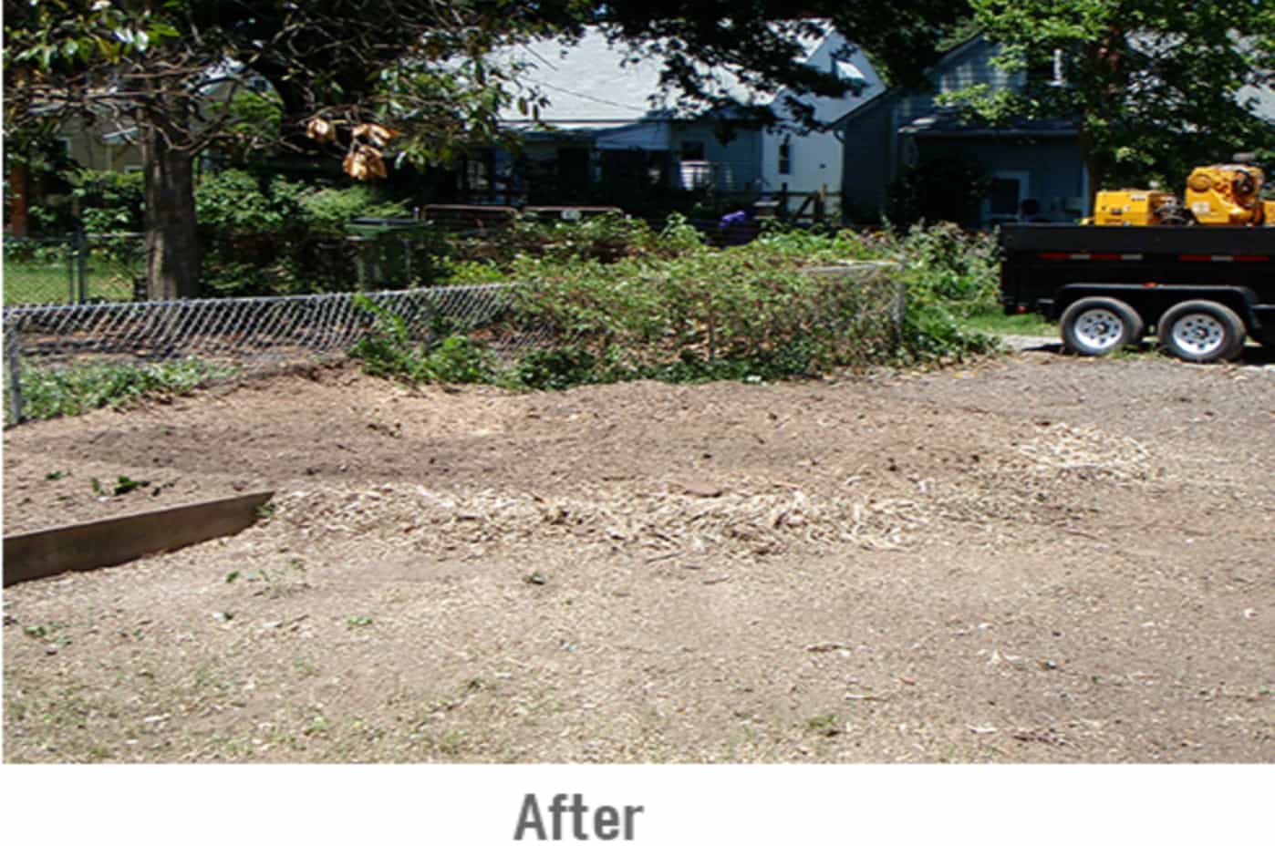 An empty dirt patch of land with a slight crater left behind after a tree stump removal.  A blue house, a truck with equipment and a large tree can be seen in the foreground