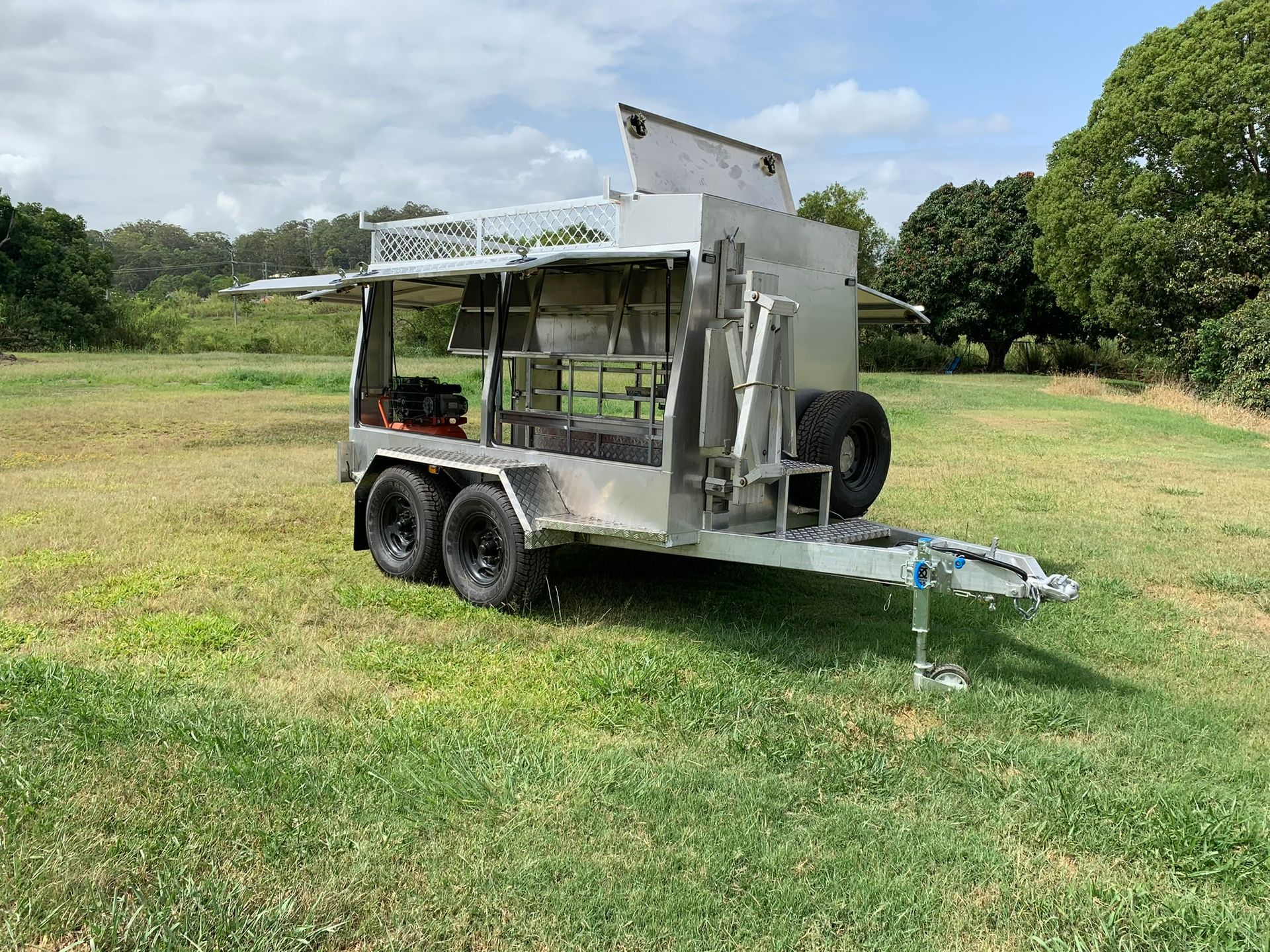 A silver, open-sided custom equipment trailer with black tires parked on a grassy field under a sunny sky — Rod's Engineering & Lifetime Trailers in Bli Bli, QLD