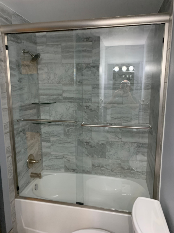 Shower stall with sliding glass doors, gray tiled walls, and a white bathtub.