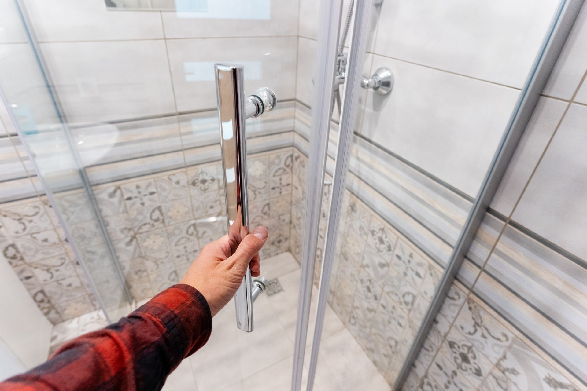 Person opening glass shower door with chrome handle in tiled bathroom.