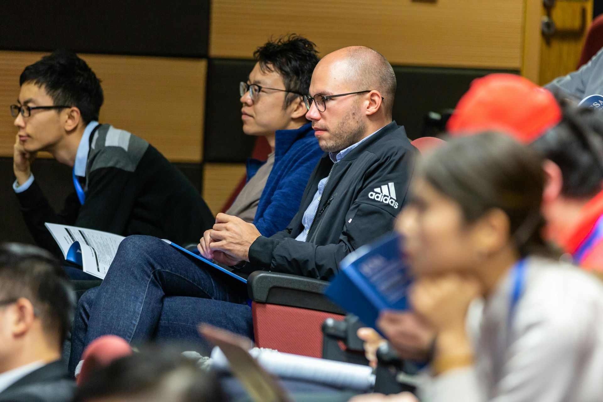 People sitting in an auditorium, listening. A man in an Adidas jacket takes notes.