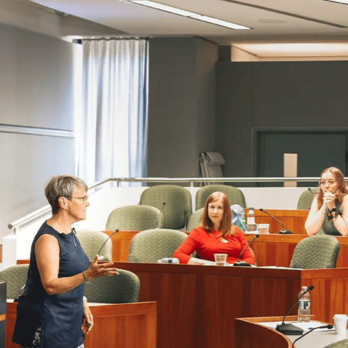 Woman speaking in a courtroom, two others seated at a table, microphones, light-colored room.
