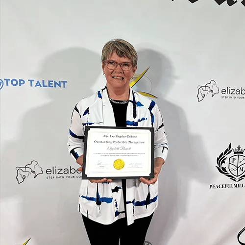 Woman with glasses holds award; white jacket with blue, black outfit; formal setting.