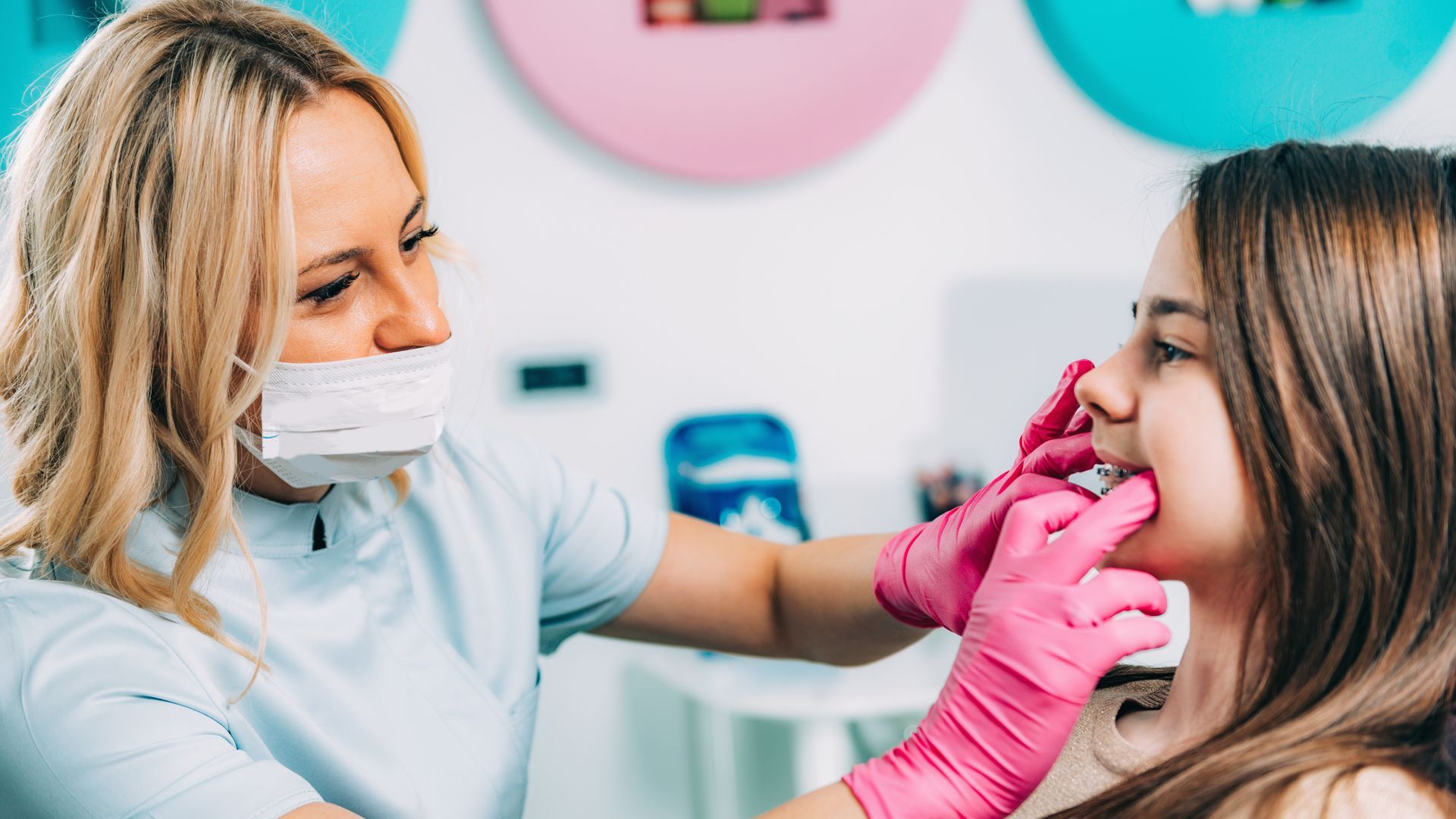 A professional in a mask and pink gloves examines a patient's teeth in a clinical setting.
