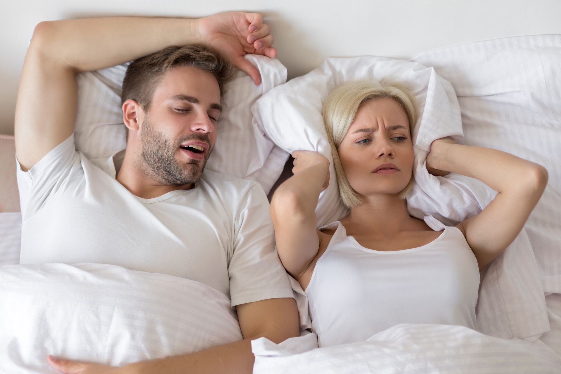 A man is snoring while a woman covers her ears in bed.