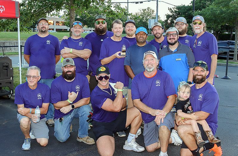 Group of men in purple shirts pose outside, some holding a trophy, smiling.