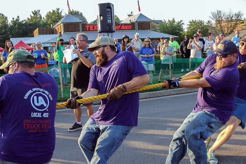 Men in purple shirts playing tug-of-war on a street, large crowd in the background, under a speaker.