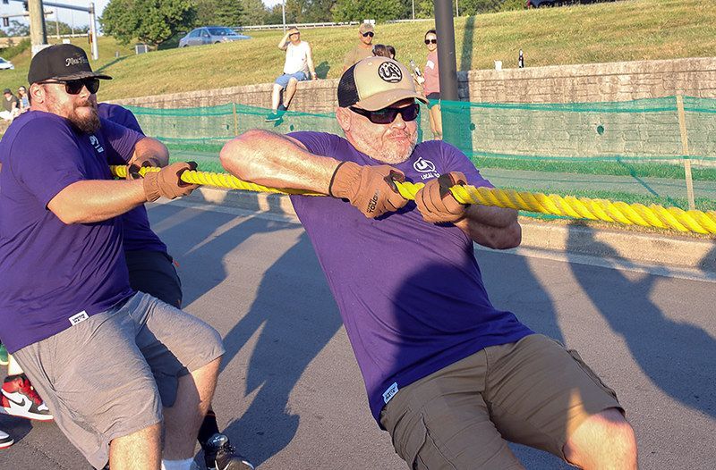 Men in purple shirts and sunglasses, pulling on a yellow rope in a tug-of-war competition outdoors.