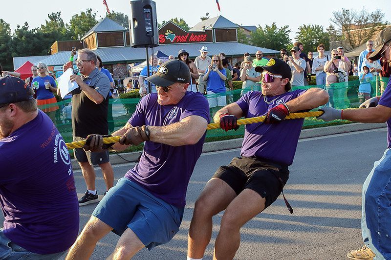 People in purple shirts pulling a rope in a tug-of-war competition on a street; crowd watches.