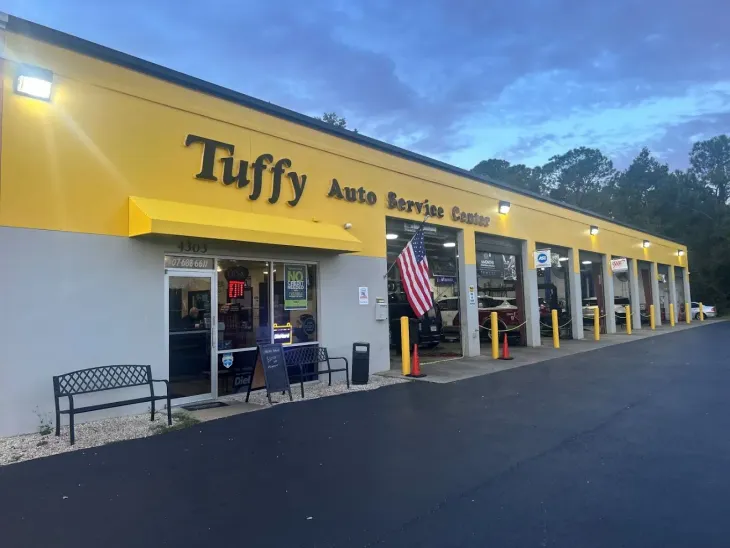 Mechanic examining a car engine in a garage. He is wearing glasses, dark clothes and is focused on the engine.
