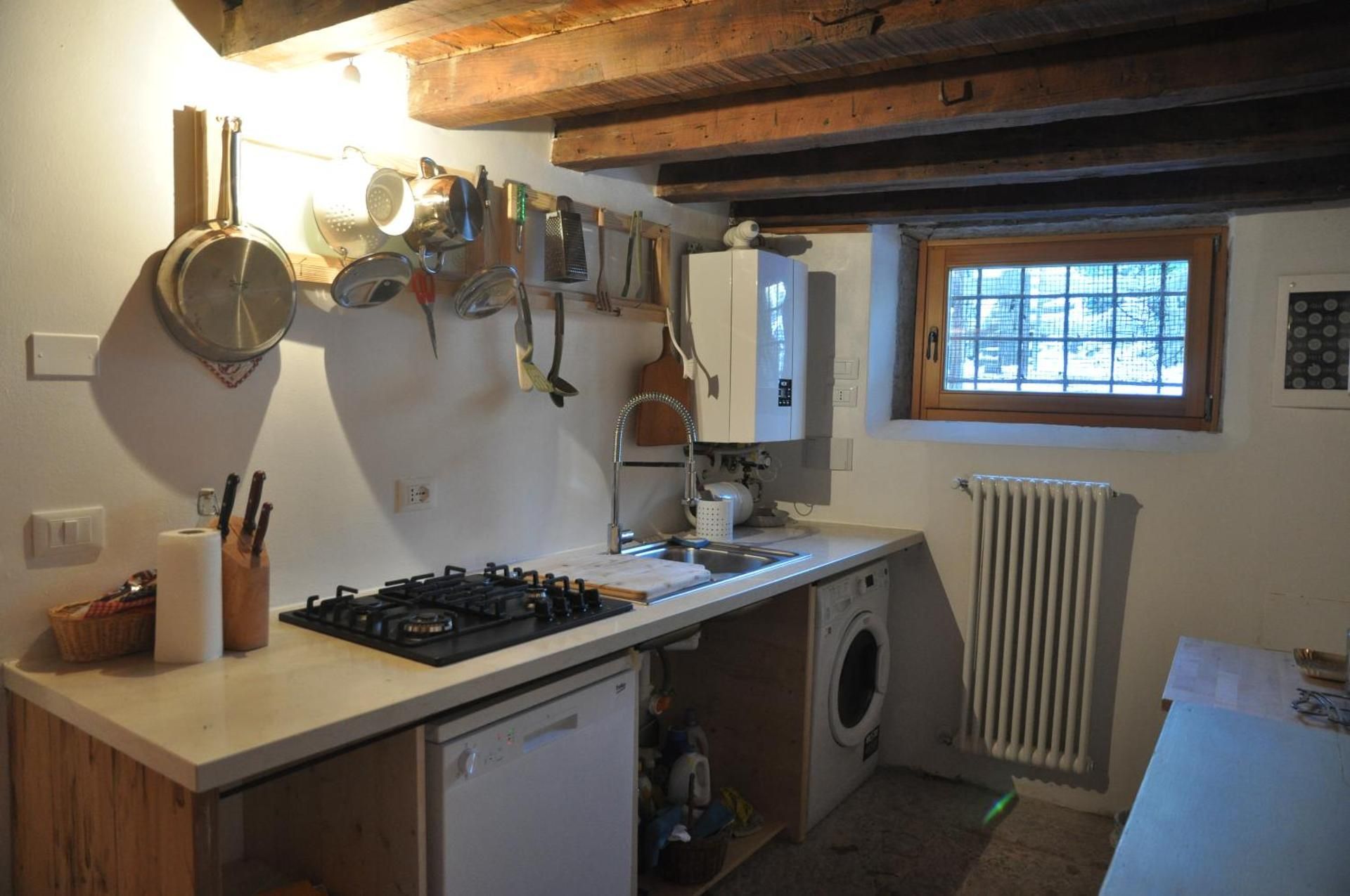 Piccola cucina rustica con elettrodomestici e pentole. Travi in ​​legno, piccola finestra, pareti bianche.