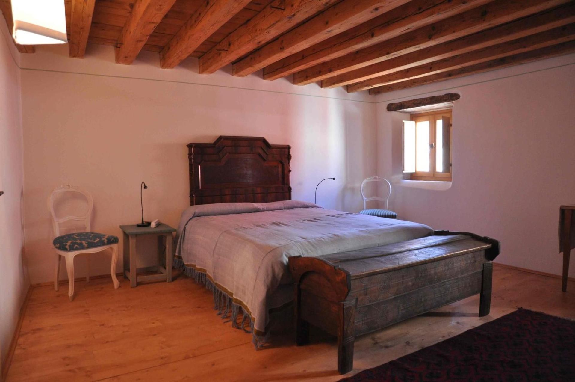 Camera da letto con letto in legno, baule, pareti bianche e soffitto con travi in ​​legno.