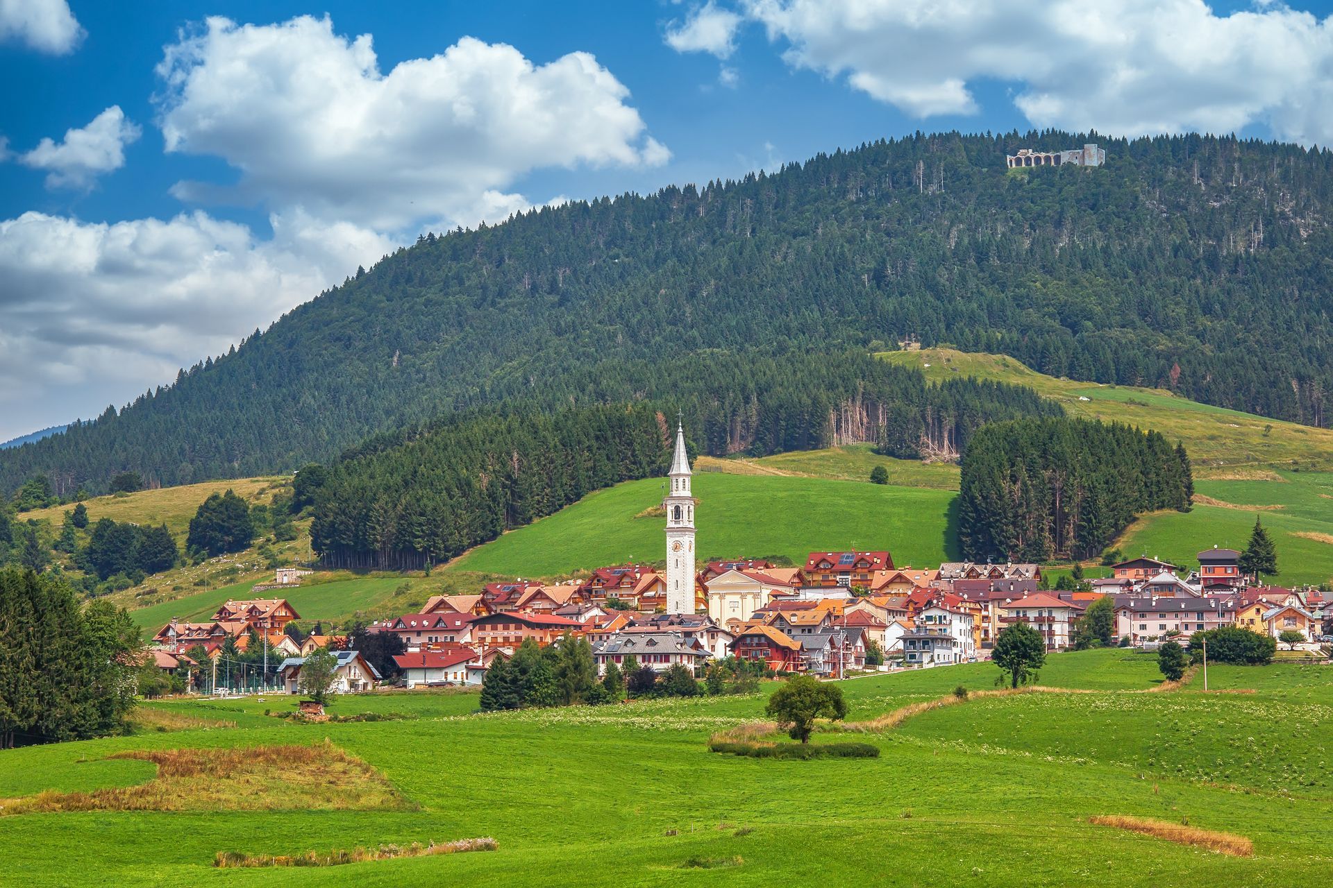 Valle verde con un villaggio e il campanile di una chiesa sullo sfondo di una montagna boscosa sotto un cielo nuvoloso.