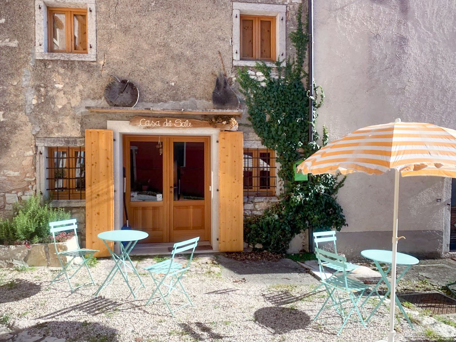 Esterno con posti a sedere all'aperto: tavoli e sedie azzurri, ombrellone a strisce arancioni, persiane e porta in legno.