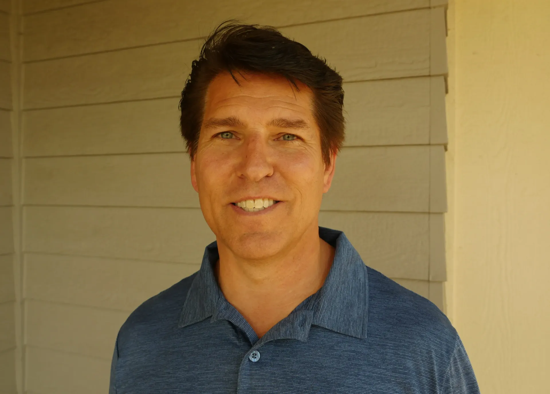A man in a blue shirt is smiling in front of a white wall.