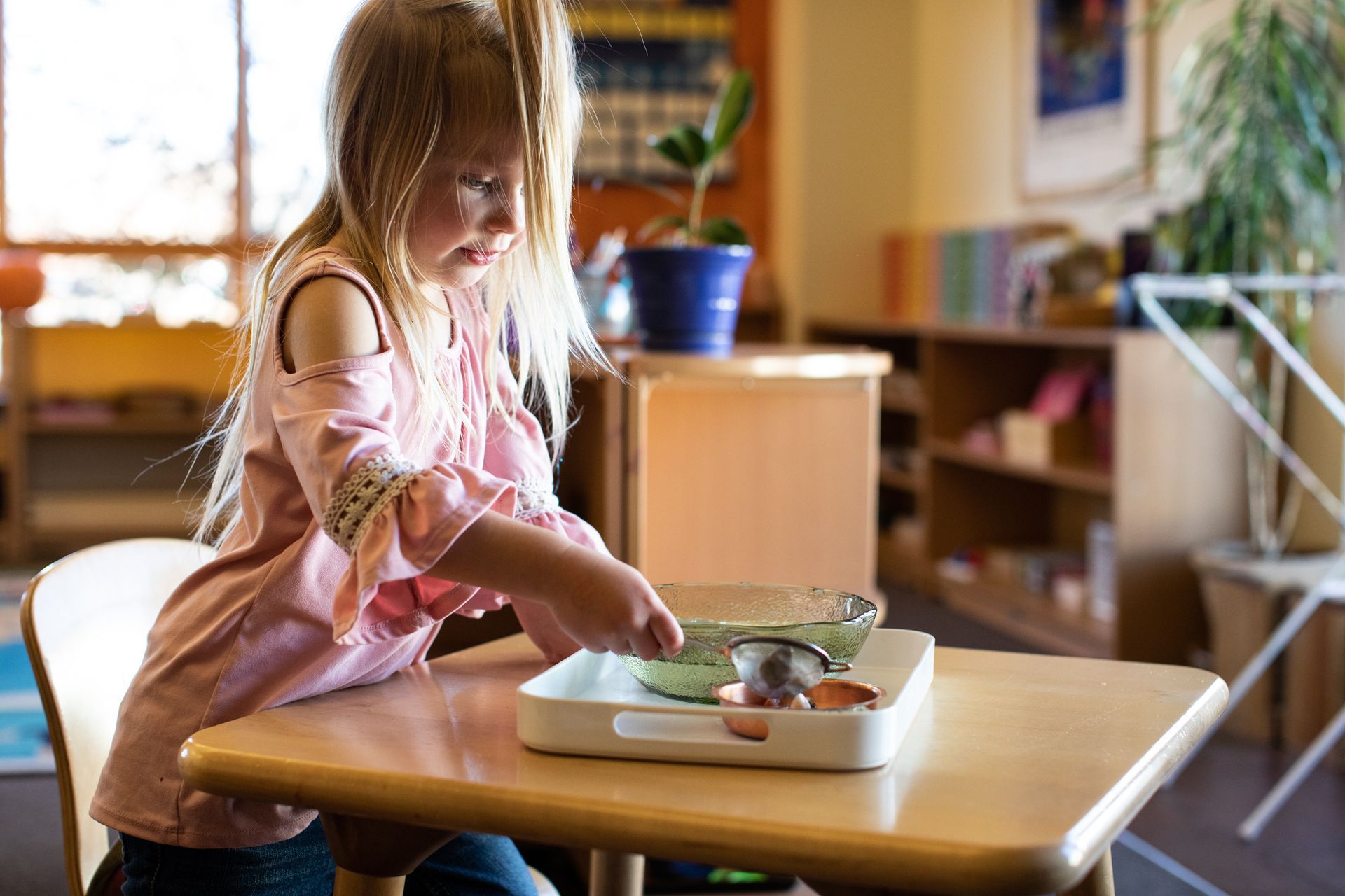 Girl at a table, playing with a bowl and strainer in a Montessori classroom.